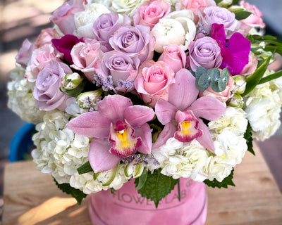 A large floral arrangement with a variety of flowers including roses and orchids, in soft colors of pink, white, and purple, presented in a round pink box.